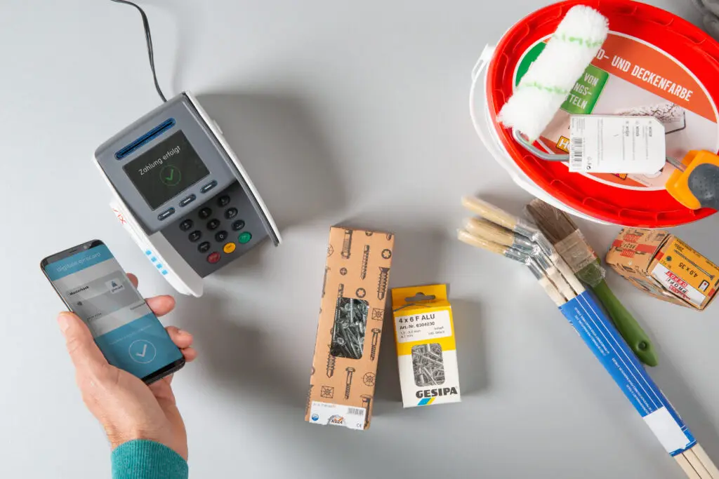 Zahlungsvorgang mit Smartphone und Kartenzahlungsgerät, umgeben von Malerzubehör wie Farbroller, Pinsel und Schraubenverpackungen auf einem grauen Tisch.