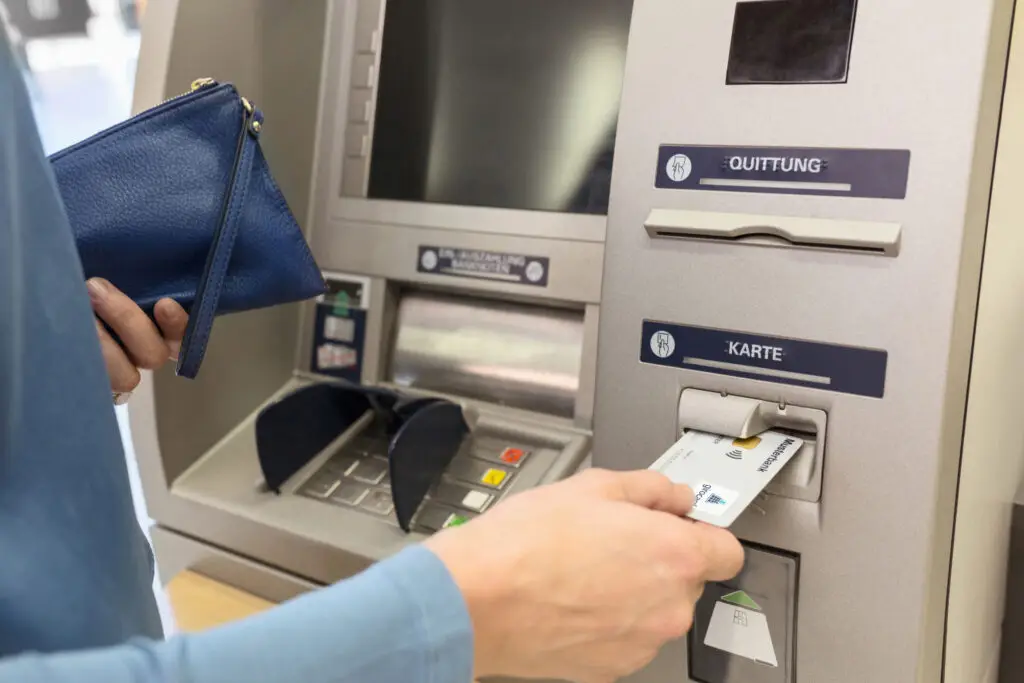 Person, die an einem Geldautomaten eine Karte einführt, während sie eine blaue Geldbörse in der anderen Hand hält. Der Bildschirm des Geldautomaten ist sichtbar, und die Tasten sind gut erkennbar.