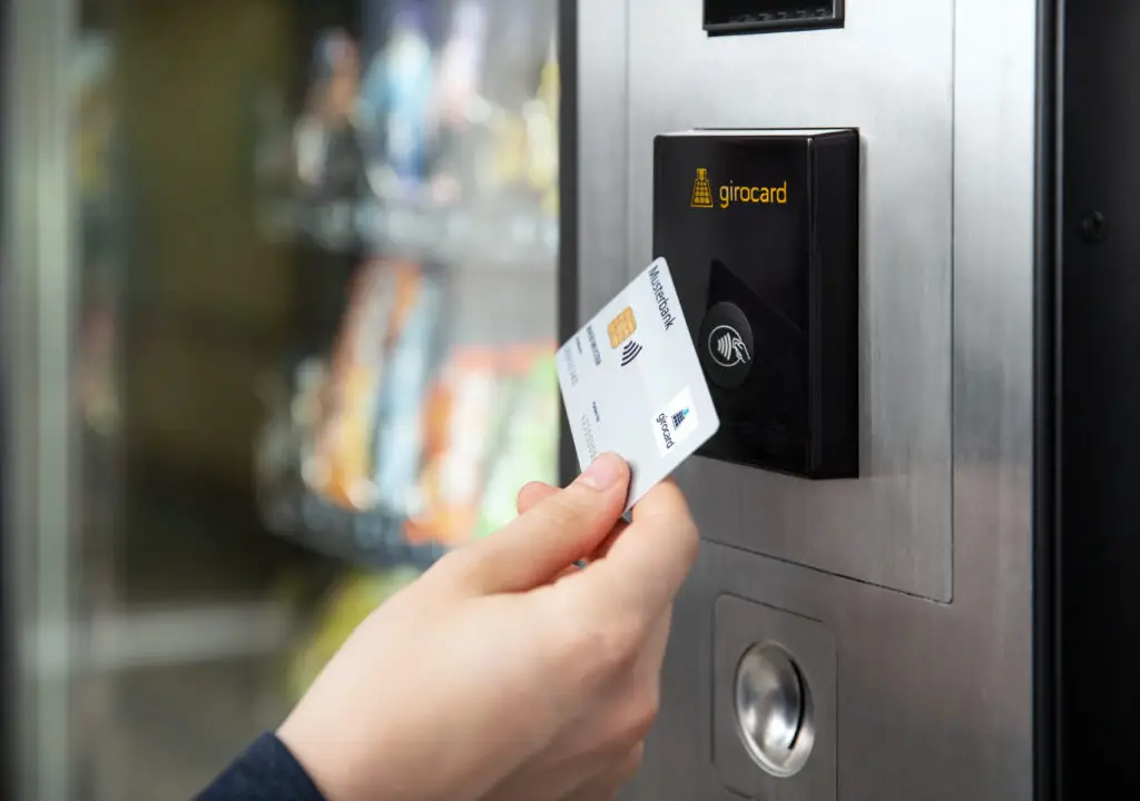 Hand, die eine girocard an einem modernen Snackautomaten vor einem Hintergrund mit Snacks hält, um eine bargeldlose Zahlung durchzuführen.