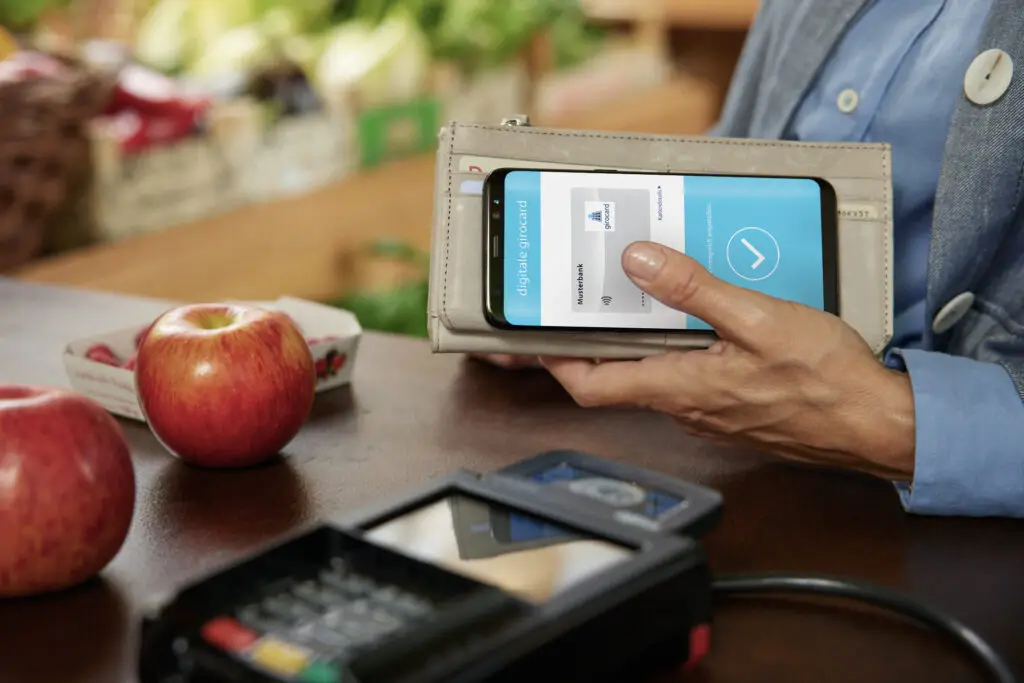 Eine Person nutzt ihr Smartphone, um eine digitale Geldbörse an einem Kassenterminal zu verwenden, während frische Äpfel und Obst im Hintergrund sichtbar sind.
