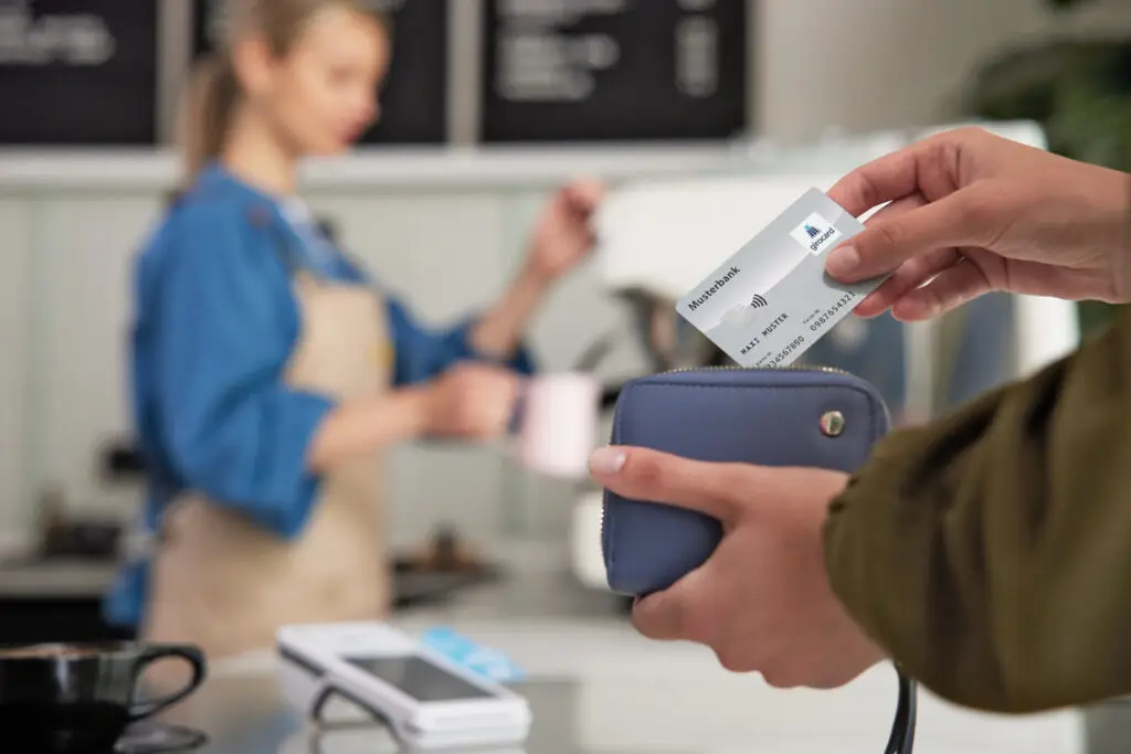 Eine Person hält eine Kreditkarte über eine Geldbörse, während im Hintergrund eine Barista einen Kaffee zubereitet. Das Bild zeigt den modernen Zahlungsprozess in einem Café.