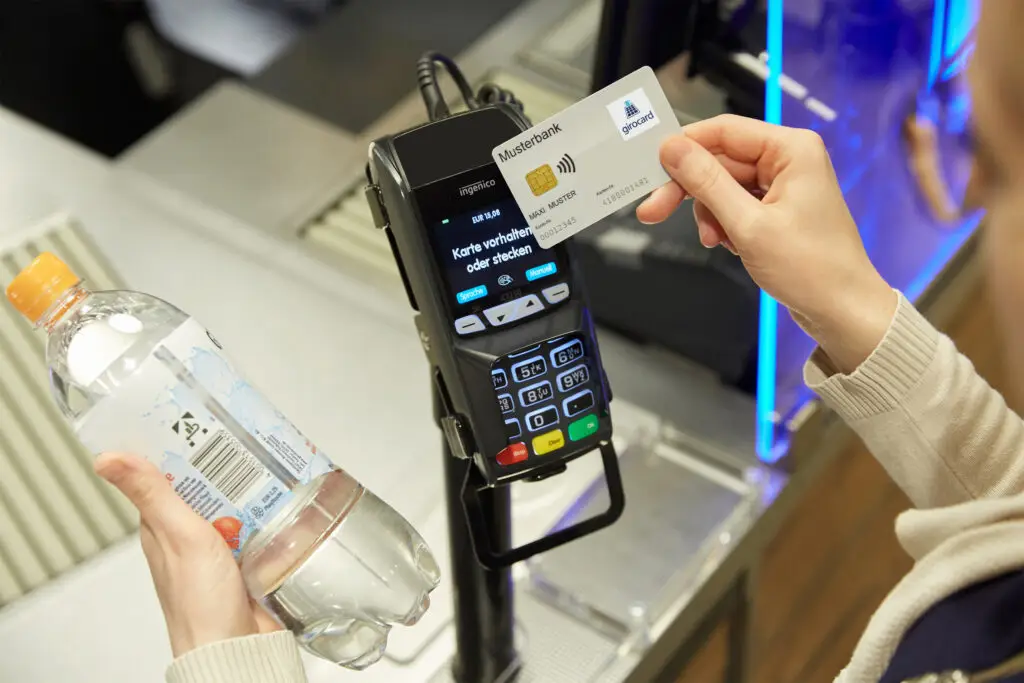 Eine Person hält eine Flasche Wasser in der linken Hand und führt eine Zahlung mit einer girocard an einem modernen Kartenzahlungsgerät durch. Das Display des Geräts zeigt Anweisungen zur Karteneinführung oder -haltung.