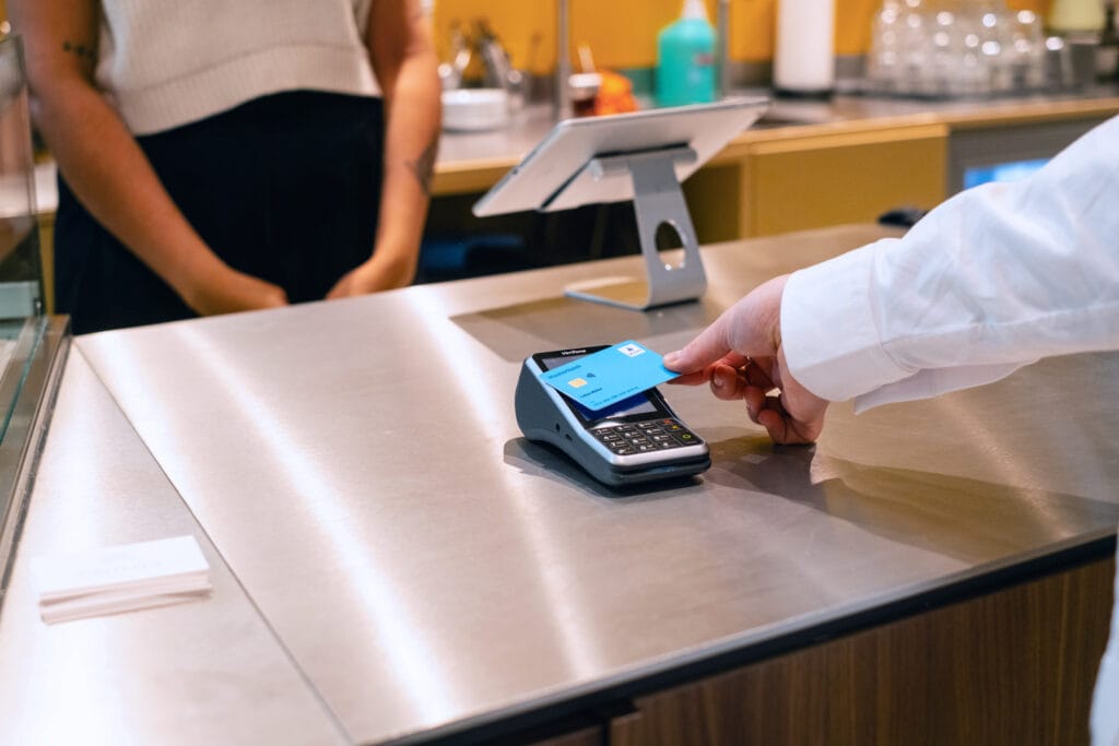 Zahlungsvorgang an der Kasse mit einem Kartenlesegerät in einem modernen Café, während eine Person eine Kreditkarte vor das Gerät hält. Im Hintergrund ist eine weitere Person zu sehen, die auf den Zahlungsvorgang wartet.