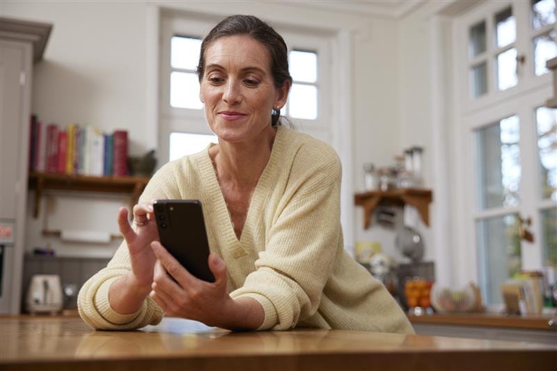 Frau in bequemer Kleidung sitzt an einem Küchentisch und schaut auf ihr Smartphone, während sie lächelt. Im Hintergrund sind Bücherregale und Küchenutensilien sichtbar.